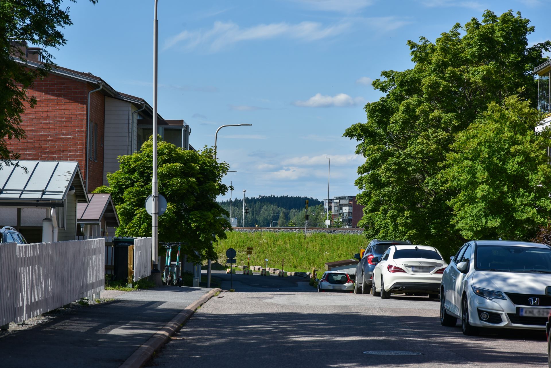 Tie, jonka takana näkyy junarata. Kadun varrella autoja, rakennuksia ja puita. 